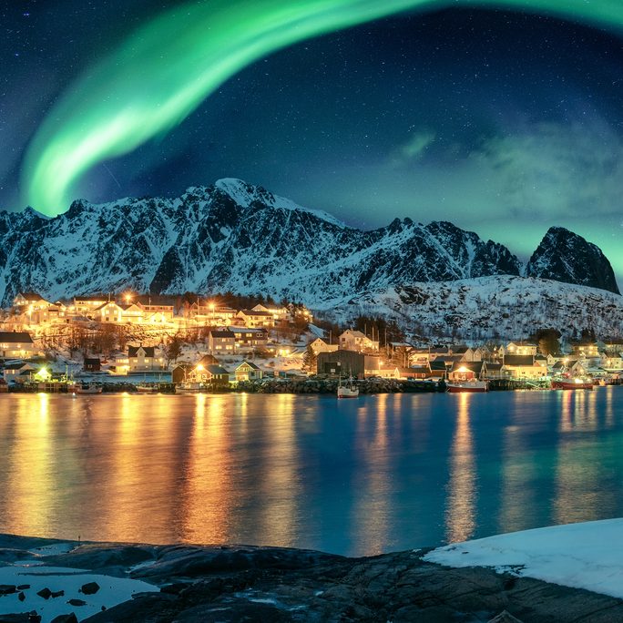 Northern lights above a lit fishing village and snow-covered peaks